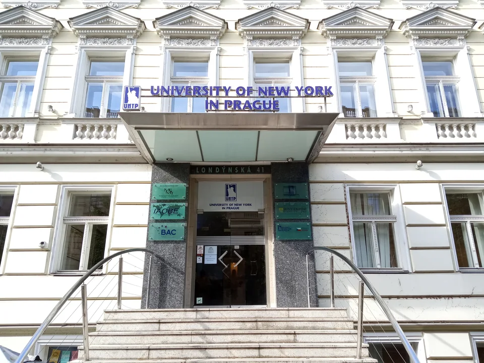 Белый исторический фасад со стеклянным козырьком и вывеской University of New York in Prague над входом, к дверям ведут ступени с металлическими перилами